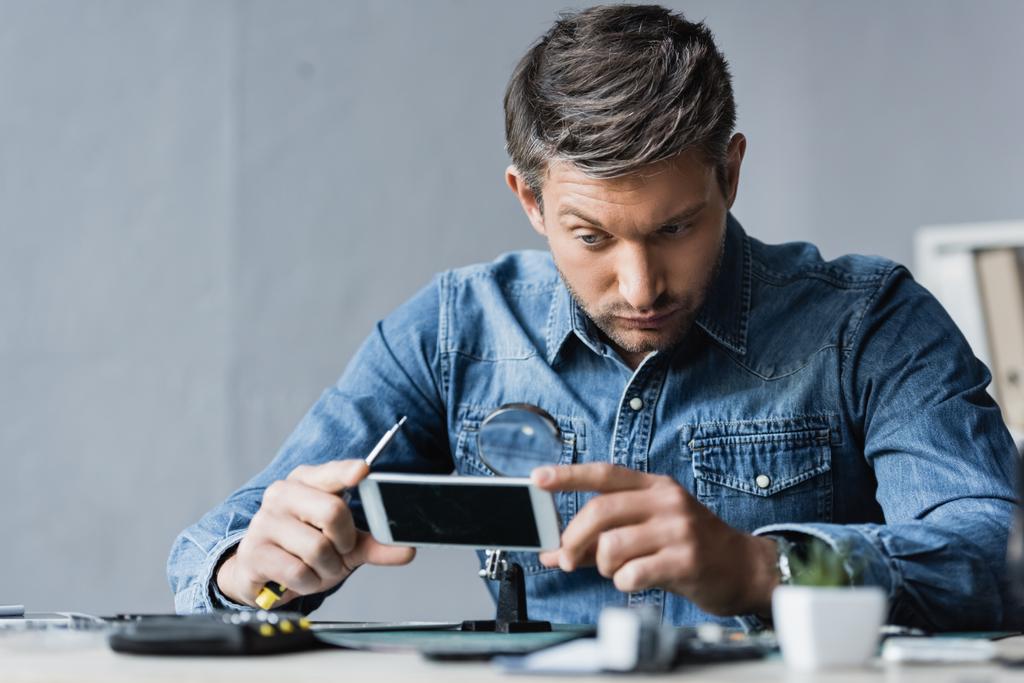 Сосредоточенный ремонтник с отверткой, смотрящий через лупу на мобильный телефон, сидя на рабочем месте на размытом переднем плане - Фото, изображение