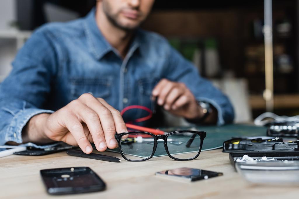 Ausgeschnittene Ansicht eines Reparateurs, der eine Brille vom Tisch nimmt, mit zerlegten Teilen von Mobiltelefonen auf verschwommenem Hintergrund - Foto, Bild