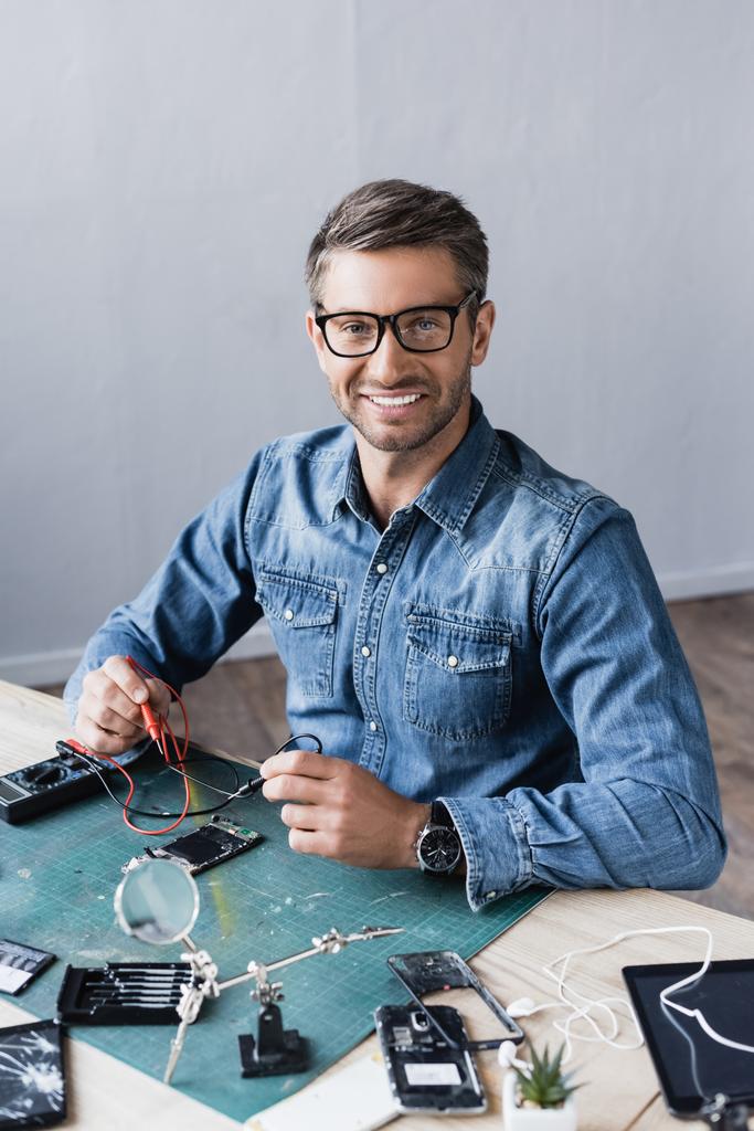 Счастливый ремонтник смотрит на камеру с сенсорами мультиметра, сидя за столом с разобранными частями смартфонов - Фото, изображение