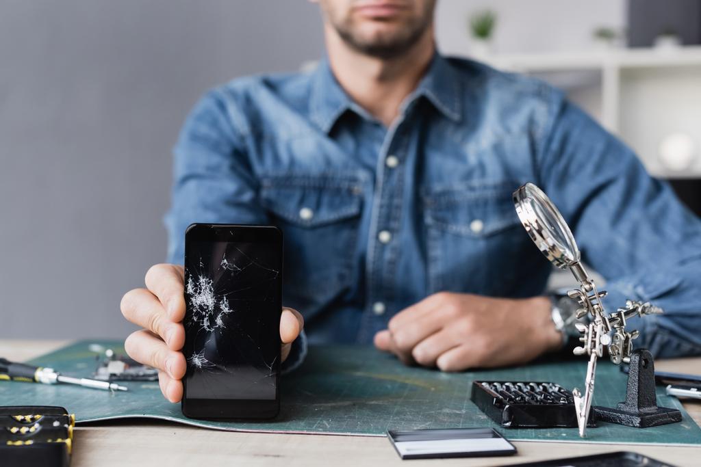 Περικοπή προβολής του επισκευαστή που δείχνει σπασμένο smartphone ενώ κάθεται στο τραπέζι με μεγεθυντικό φακό σε θολή φόντο - Φωτογραφία, εικόνα
