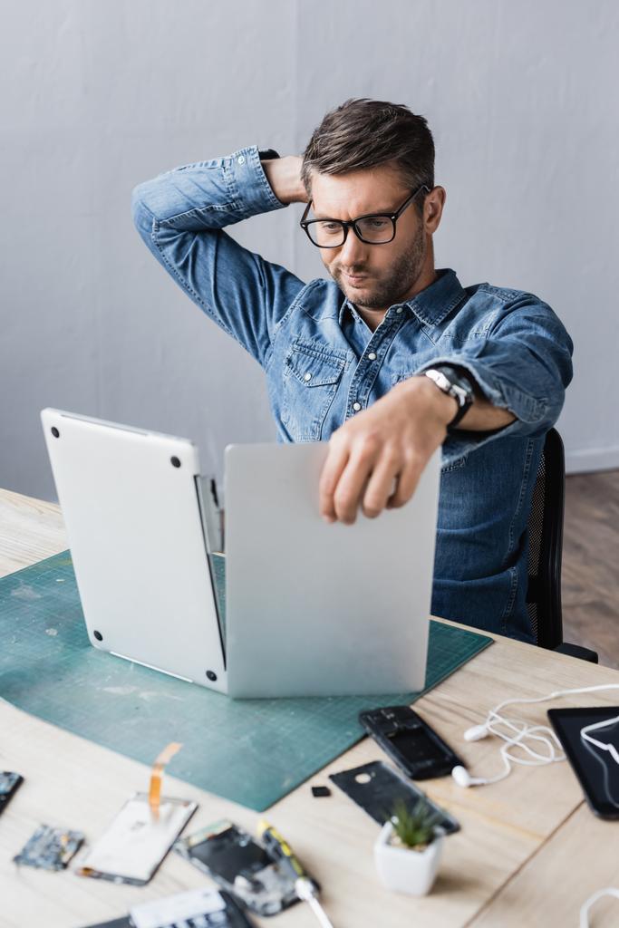 Задумчивый ремонтник с затылком смотрит на сломанный ноутбук на рабочем месте на размытом переднем плане - Фото, изображение