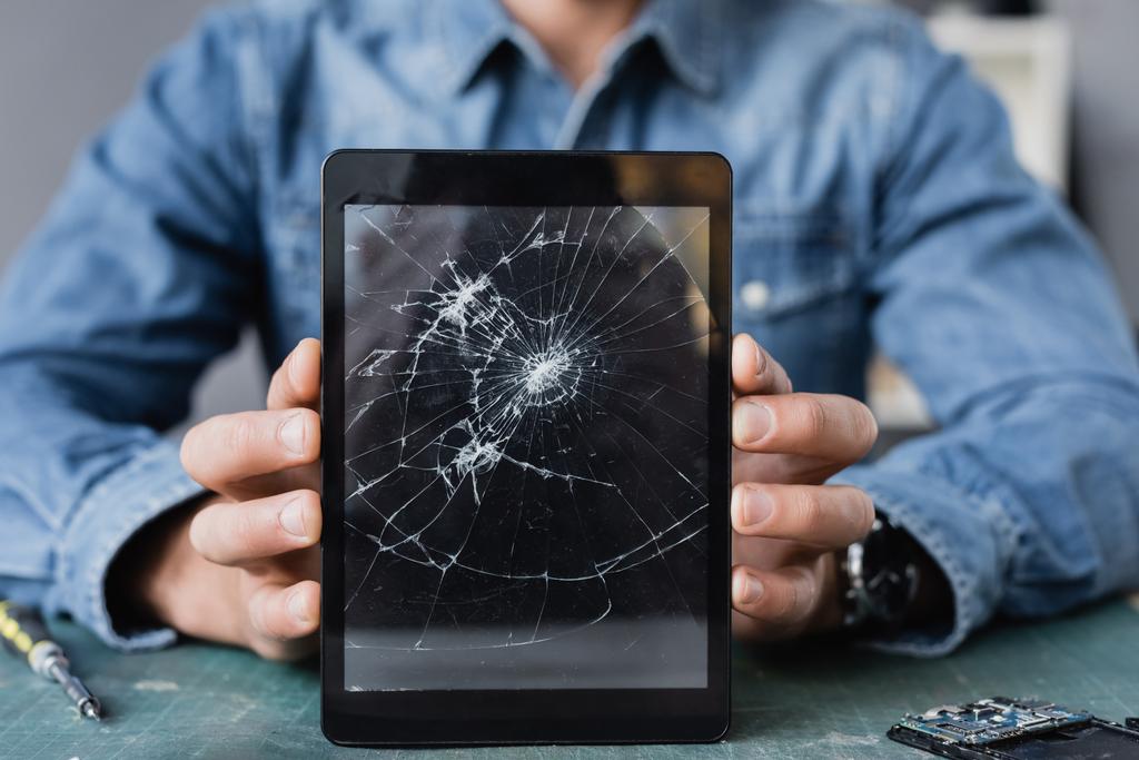 Nahaufnahme eines zertrümmerten digitalen Tablets in den Händen eines Reparateurs am Arbeitsplatz auf verschwommenem Hintergrund - Foto, Bild