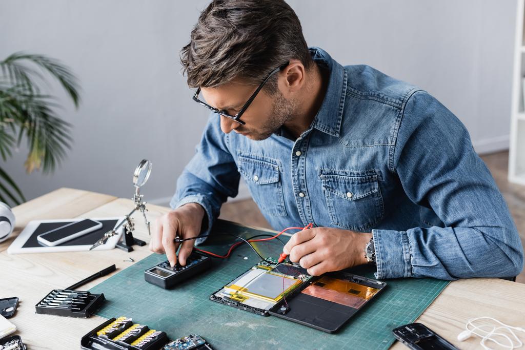 Reparador regulando multímetro enquanto segurando sensores perto de parte desmontada de tablet digital quebrado no local de trabalho - Foto, Imagem