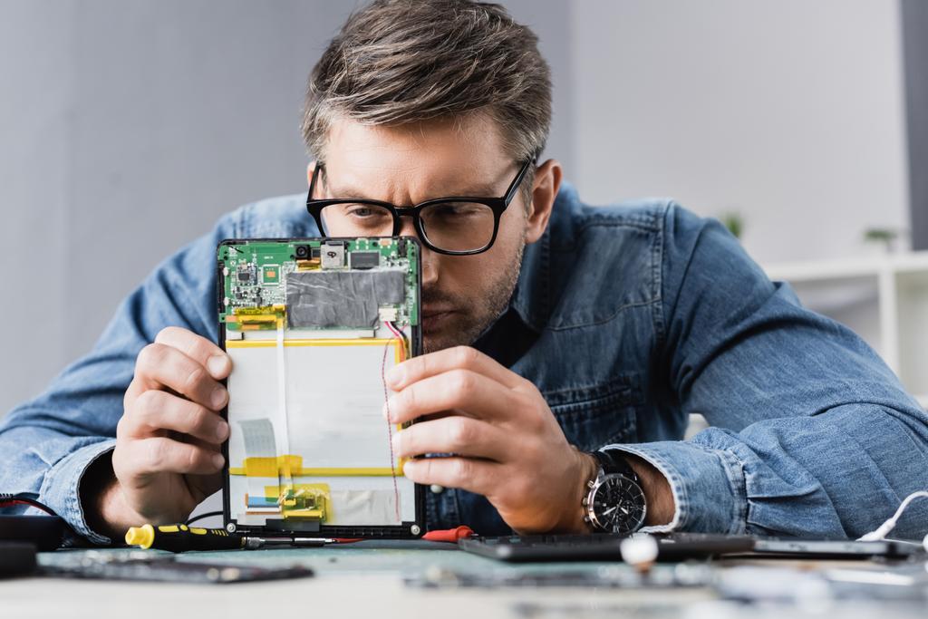 Konzentriertes Schielen des Reparateurs, während er einen Teil des kaputten digitalen Tablets mit verschwommenem Arbeitsplatz im Vordergrund betrachtet - Foto, Bild