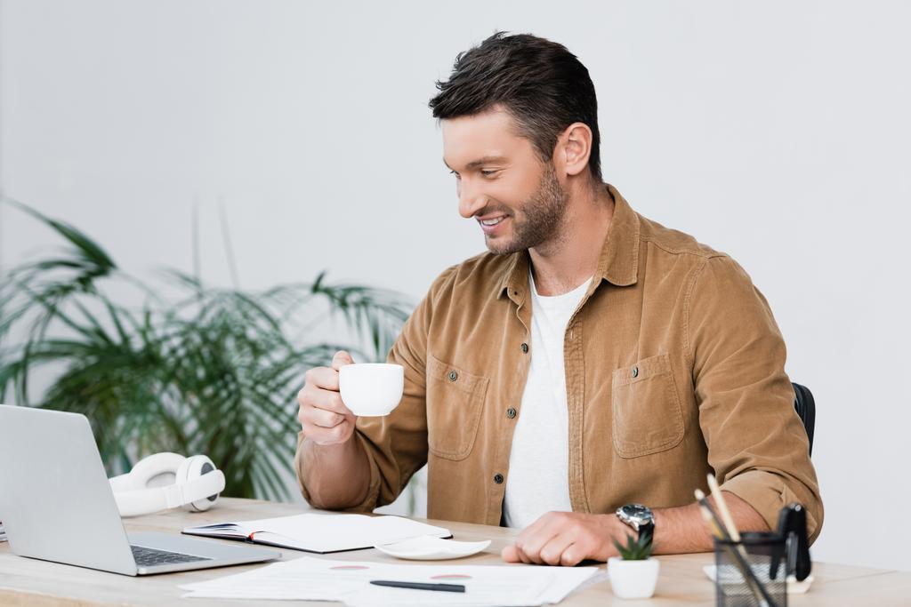 Vrolijke zakenman met koffiekop op zoek naar laptop tijdens het zitten op het werk met wazig plant op de achtergrond - Foto, afbeelding