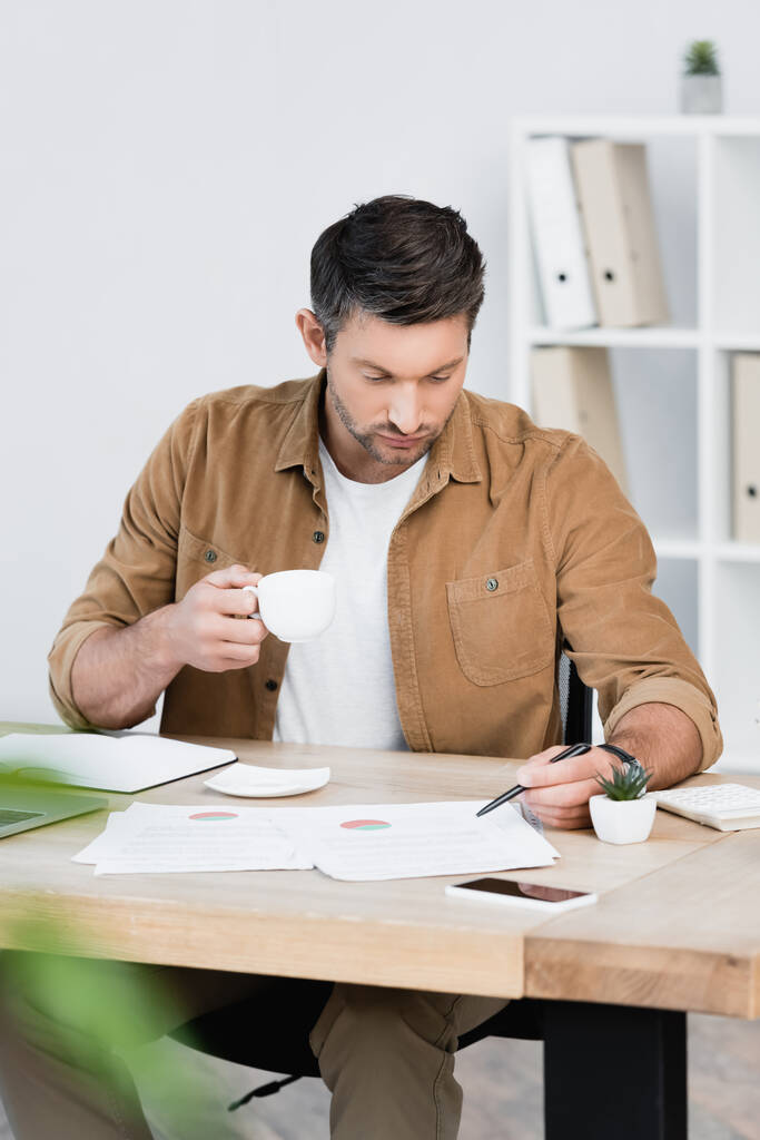 Hombre de negocios enfocado con taza de café apuntando con bolígrafo en la hoja de papel mientras está sentado en el lugar de trabajo en primer plano borroso - Foto, imagen