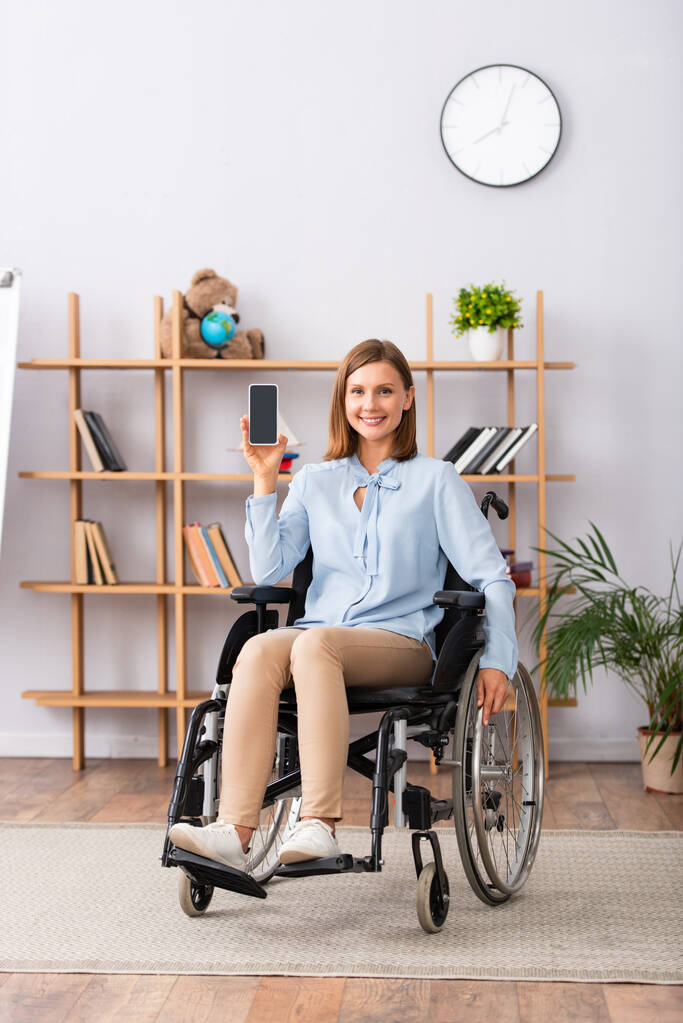 Longitud completa de una psicóloga sonriente que muestra un teléfono inteligente con pantalla en blanco mientras está sentada en silla de ruedas en la oficina - Foto, Imagen