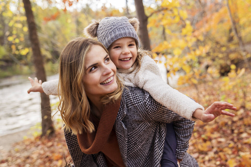 Κοριτσάκι και η μητέρα της παίζουν στο πάρκο το φθινόπωρο με το παιδί στην πλάτη - Φωτογραφία, εικόνα