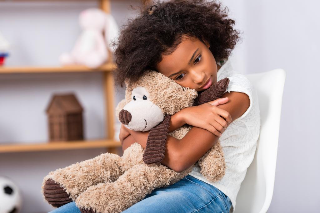 Depresso africano americano ragazza con autismo abbracciare orsacchiotto mentre seduto sulla sedia con ufficio offuscata su sfondo - Foto, immagini