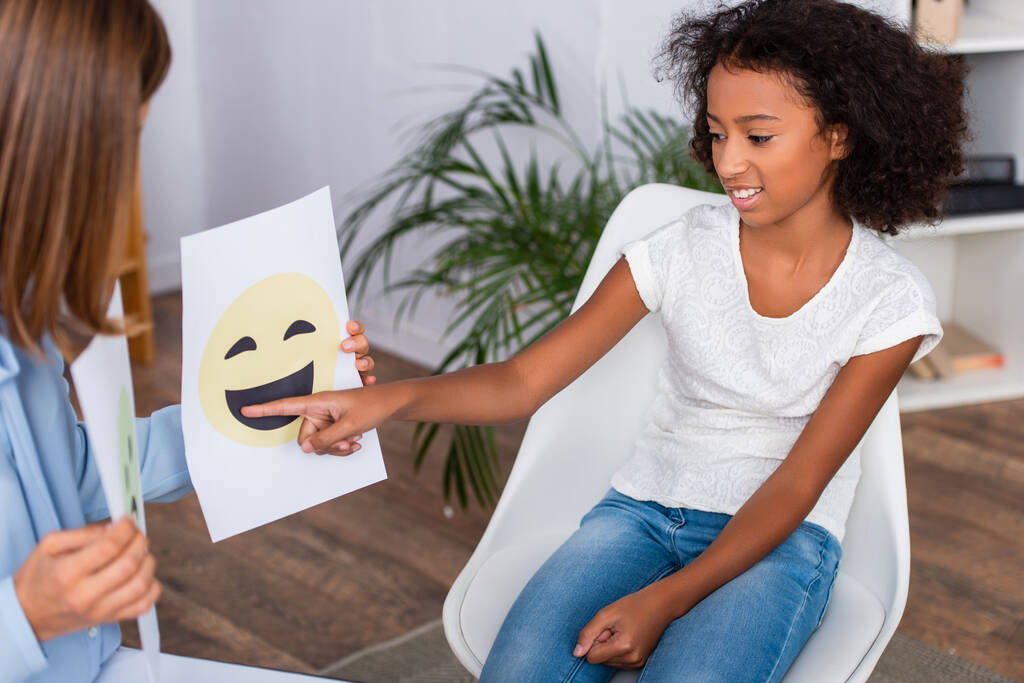 Uśmiechnięta Afroamerykanka wskazująca palcem na szczęśliwy wyraz twarzy na papierze w pobliżu psychologa na niewyraźnym pierwszym planie - Zdjęcie, obraz