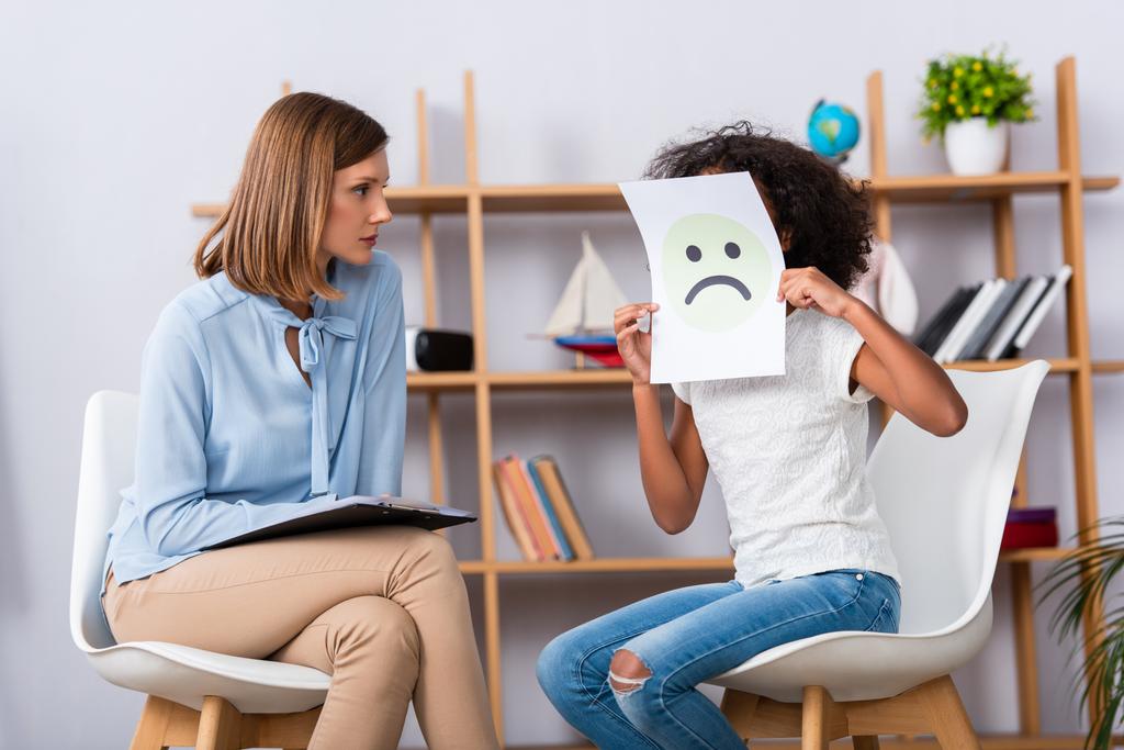 Psychologist looking at african american girl covering face with unhappy expression on paper with blurred office on background - Photo, Image