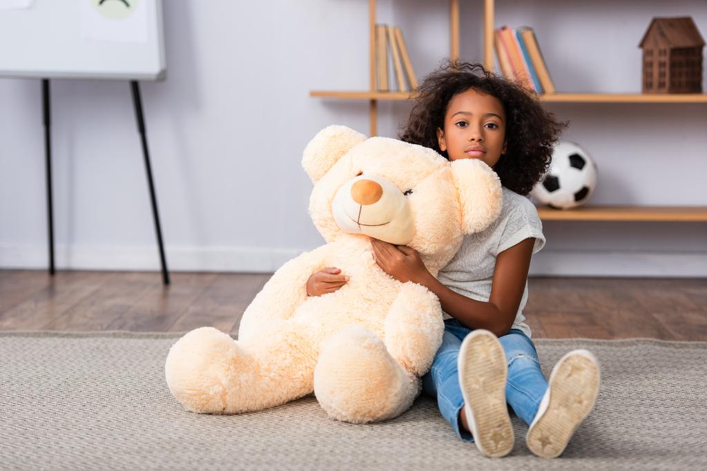 Afectada chica afroamericana con autismo mirando a la cámara y abrazando oso de peluche mientras está sentado en el suelo con la oficina borrosa en el fondo - Foto, imagen