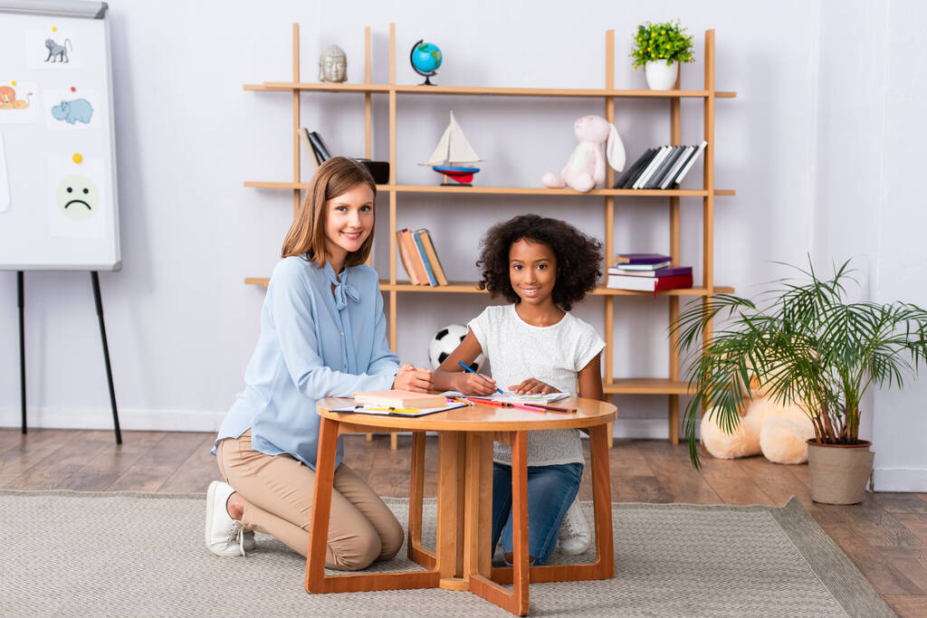 Full length of happy multicultural psychologist and girl looking at camera while sitting near coffee table in office - Photo, Image