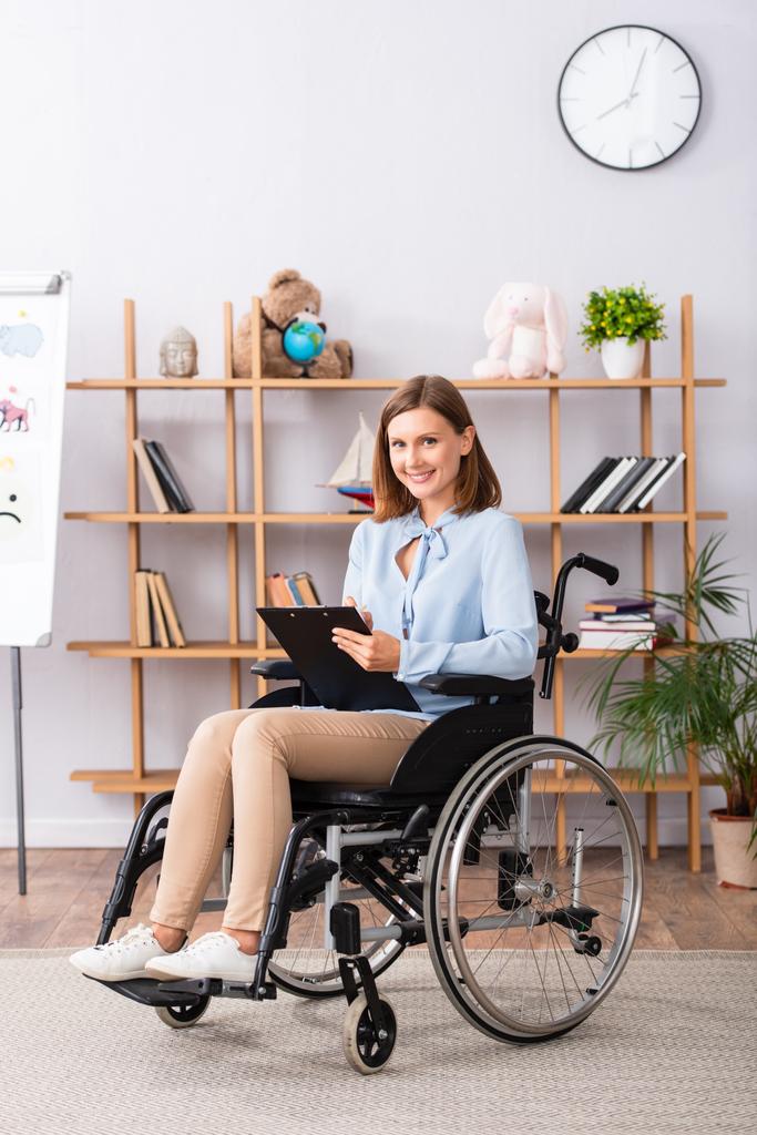 Volledige lengte van de glimlachende psycholoog met klembord kijken naar de camera tijdens het zitten in een rolstoel op kantoor - Foto, afbeelding