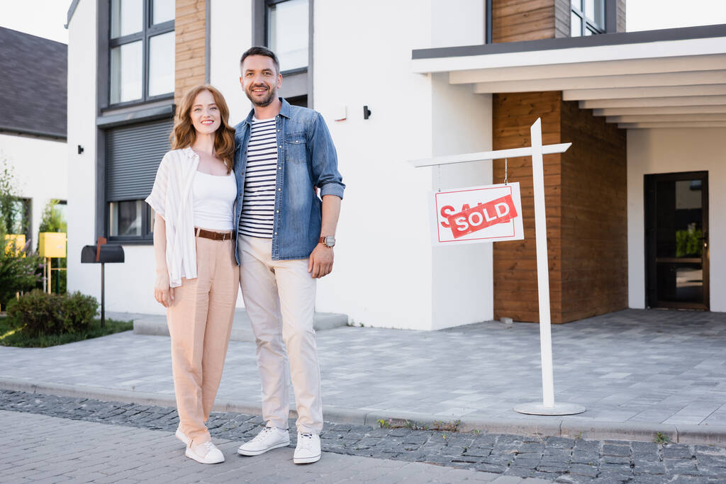 Pełna długość szczęśliwej pary patrzącej w kamerę i przytulającej się stojąc przy znaku ze sprzedawanymi literami i domem - Zdjęcie, obraz