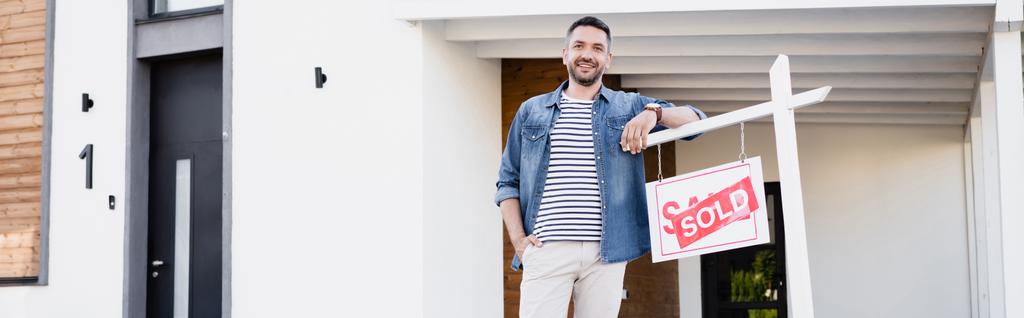 Happy man with hand in pocket looking at camera while leaning on sign with sold lettering, banner - Photo, Image