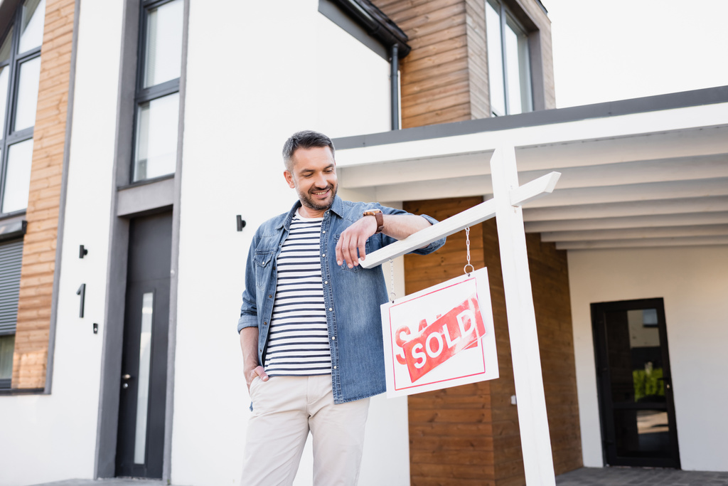 Uśmiechnięty mężczyzna z ręką w kieszeni patrząc na znak ze sprzedawanym napisem w pobliżu domu - Zdjęcie, obraz