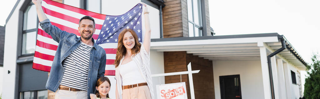 Glückliches Paar mit Tochter, die in die Kamera schaut, während sie die amerikanische Flagge in der Nähe des Schildes mit verkauftem Schriftzug und Haus hält, Banner - Foto, Bild
