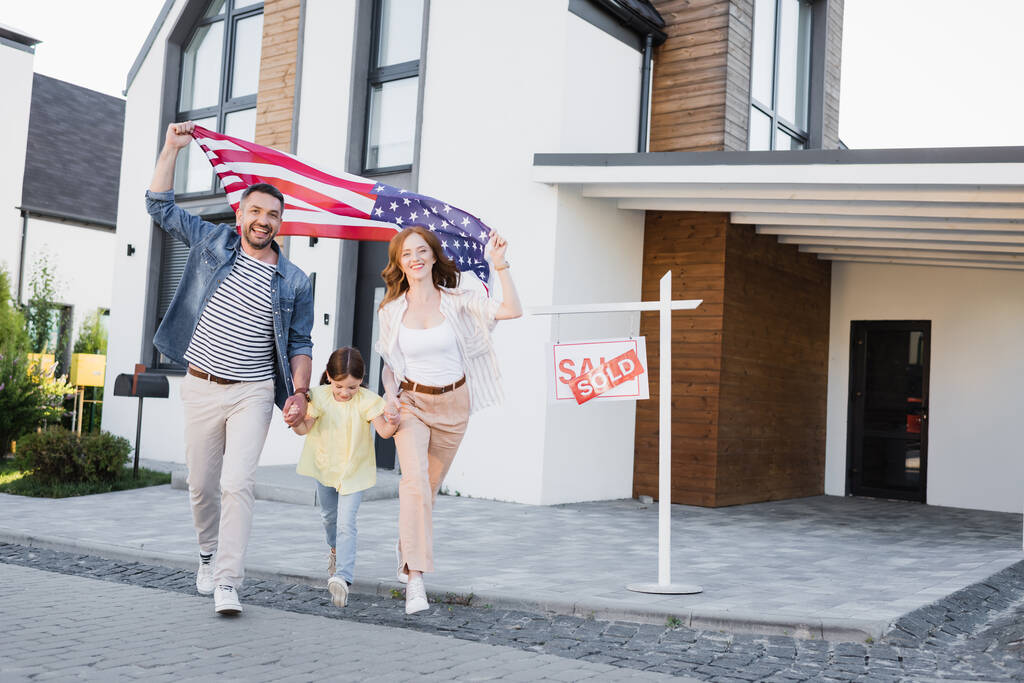 Daughter with happy mom and dad holding american flag going forward near sign with sold lettering and house - Photo, Image