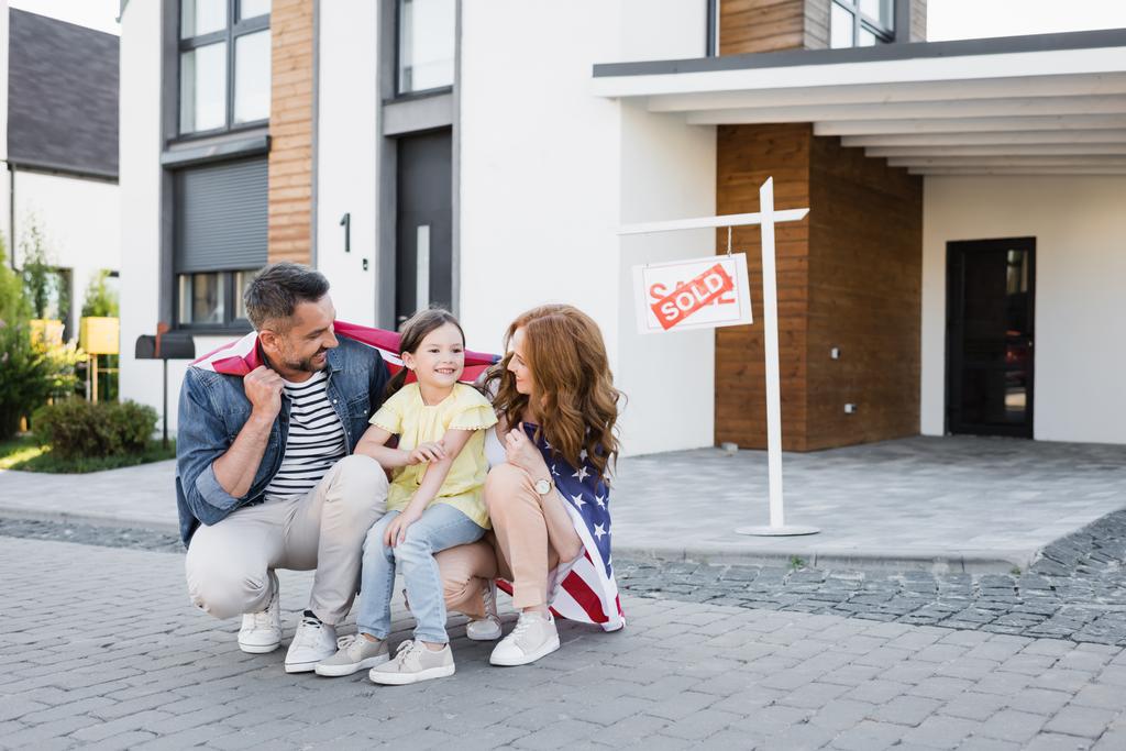 Šťastný pár s americkou vlajkou při pohledu na dceru, zatímco dřepí v blízkosti znamení s prodaným nápisem a domu - Fotografie, Obrázek