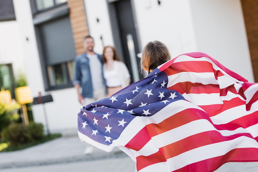 Rückansicht der Tochter mit amerikanischer Flagge und verschwommenen Eltern im Hintergrund - Foto, Bild