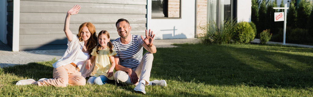 Szczęśliwi rodzice z machającymi rękami i córka siedzi na trawniku i patrząc na kamerę w pobliżu domu, sztandar - Zdjęcie, obraz