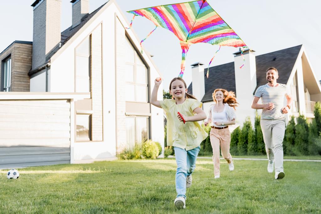 Glückliche Tochter blickt in die Kamera, während sie mit ihren Eltern im Hintergrund in der Nähe ihres Hauses Drachen fliegt - Foto, Bild