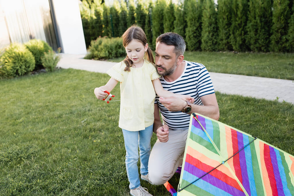 Daughter with kite string holder standing with father squatting near on lawn on blurred background - Photo, Image
