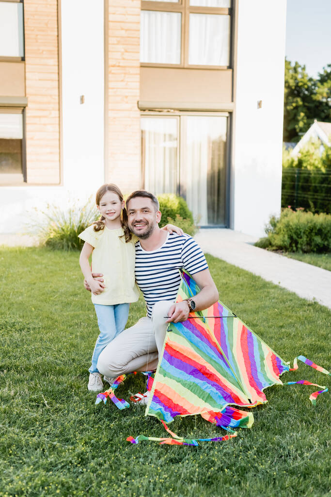 szczęśliwy ojciec z latawcem przytulanie córka i patrząc w aparat na trawniku z domu na tle - Zdjęcie, obraz