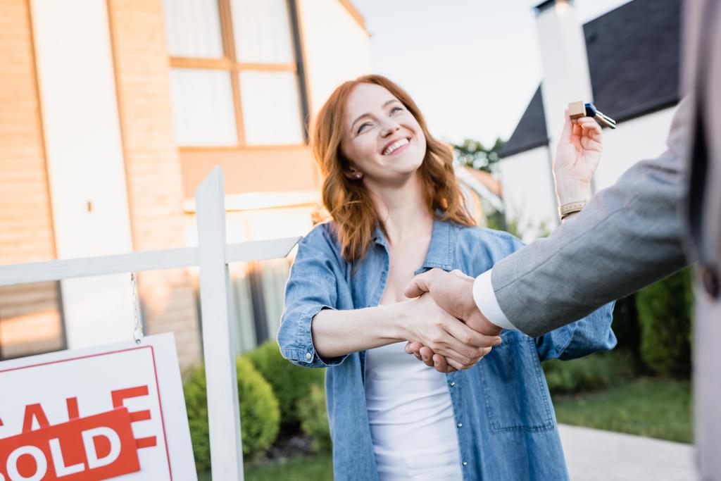 Happy redhead woman with keys shaking hands with broker near sign with blurred houses on background - Photo, Image