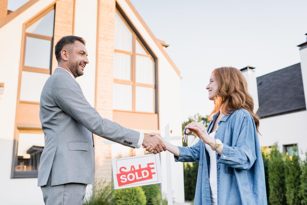 Side view of happy broker and woman with keys shaking hands near sign with sold lettering on blurred background - Photo, Image