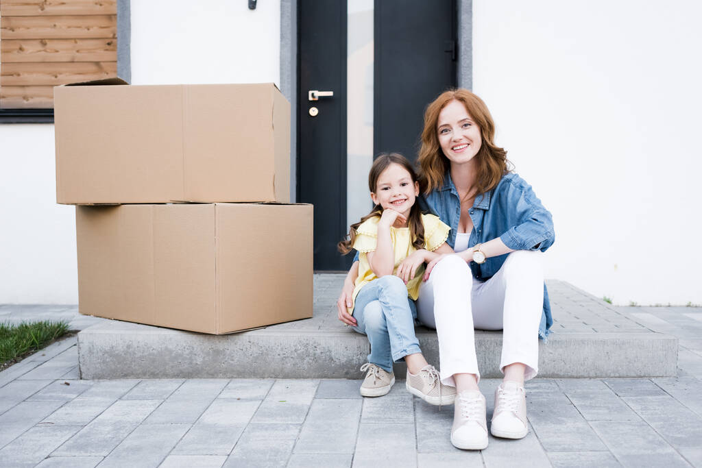 Smiling redhead mother hugging daughter while sitting on doorstep near carton boxes - Photo, Image