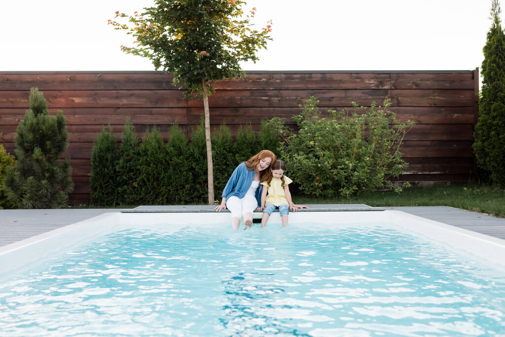 Feliz madre e hija sentadas con piernas en la piscina en el patio trasero - Foto, Imagen