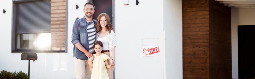 Szczęśliwa rodzina patrząc w kamerę, stojąc razem w pobliżu domu i podpisując sprzedawane litery, baner - Zdjęcie, obraz