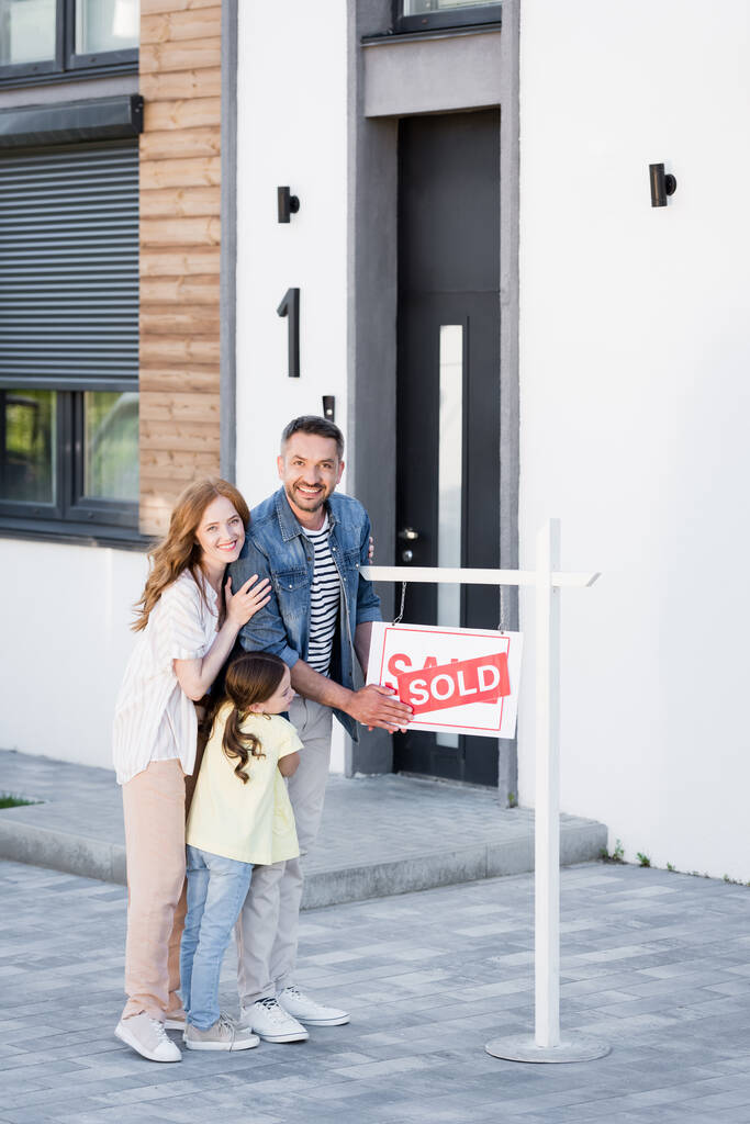 Full length of happy family hugging while looking at camera near sign with sold lettering and house - Photo, Image