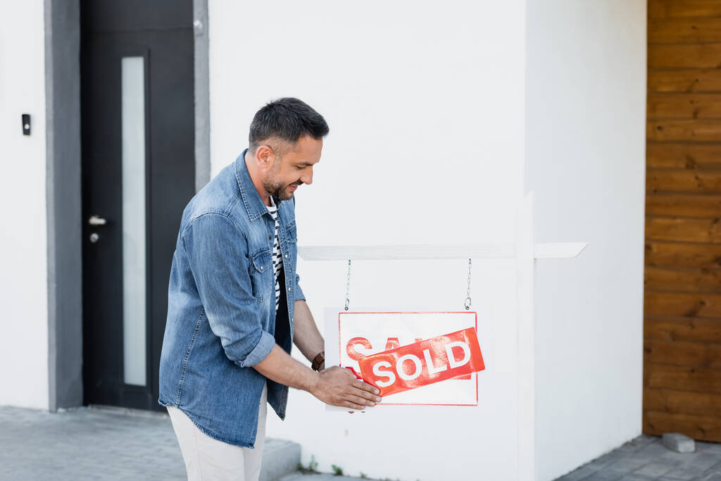 Pozytywny brodaty mężczyzna trzyma znak ze sprzedawaną literą w pobliżu domu - Zdjęcie, obraz