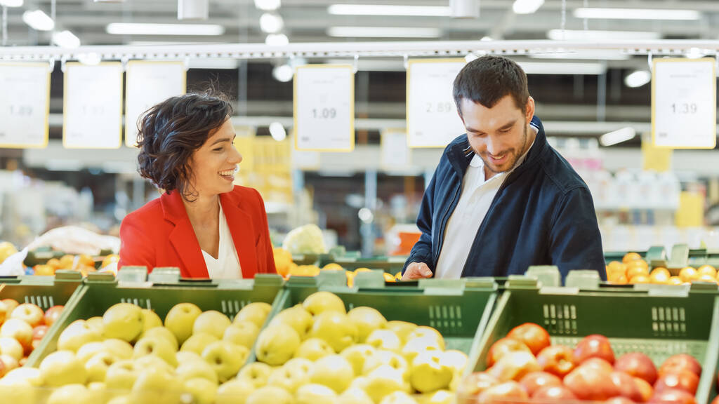 Im Supermarkt: Glückliches junges Paar entscheidet sich für Bio-Obst im Frischebereich des Ladens Ehemann nutzt Smartphone, Ehefrau holt sich Obst. - Foto, Bild