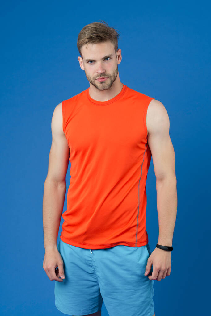 Αθλητής σε βιολετί φόντο. Μυώδης άντρας με πορτοκαλί γιλέκο και μπλε σορτσάκι. Σε φόρμα και αυτοπεποίθηση. Αθλητική μόδα για προπόνηση και προπόνηση. Προσωπικός γυμναστής για υγιεινό τρόπο ζωής. Γυμναστική και ευεξία. - Φωτογραφία, εικόνα