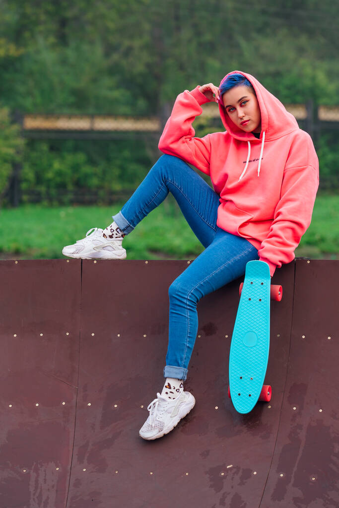 Портрет модной молодой девушки в розовой толстовке и джинсах, сидящей рядом со скейтбордом со своим голубым пластиковым скейтбордом. - Фото, изображение