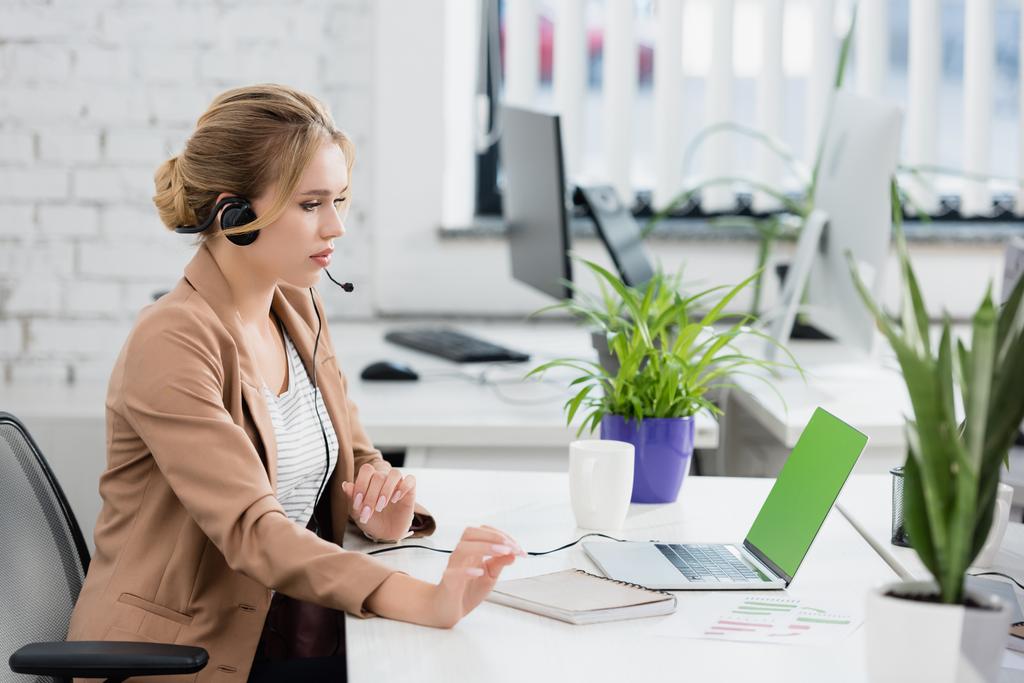 Kulaklık takmış sarışın bir kadın bilgisayarına bakarken, iş yerinde dijital cihazlarla otururken bulanık arka planda - Fotoğraf, Görsel