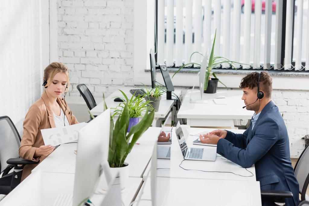 Callcenter-Mitarbeiter in Headsets arbeiten, während sie im Büro an Tischen mit digitalen Geräten sitzen  - Foto, Bild