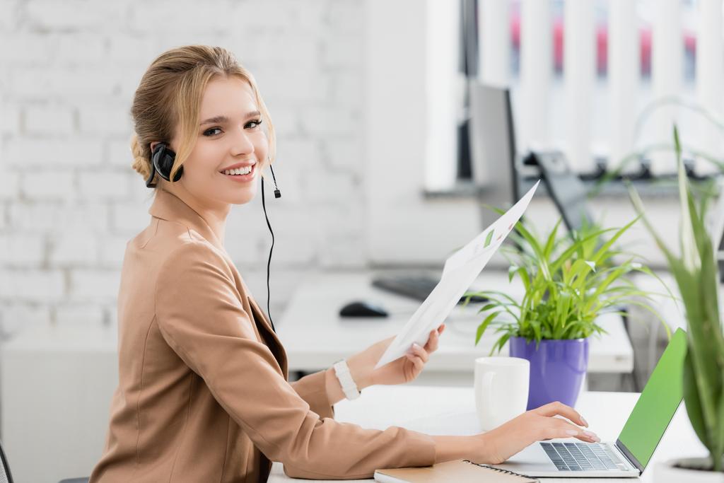 Mujer sonriente en auriculares con hoja de papel mirando a la cámara, mientras está sentada en el lugar de trabajo con el ordenador portátil en un fondo borroso - Foto, Imagen