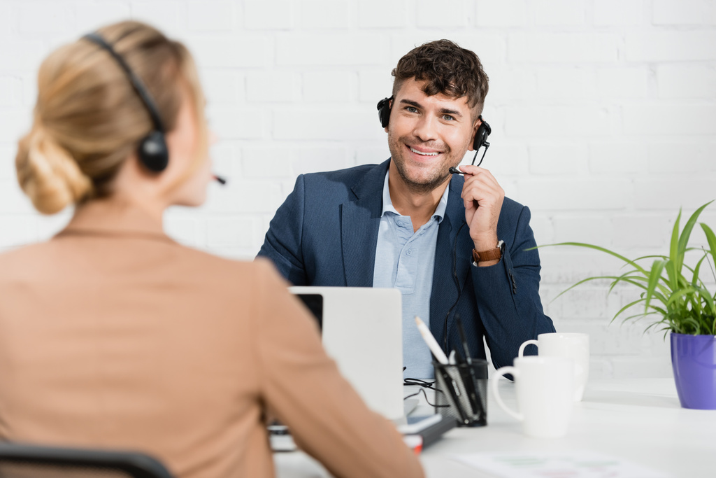 Operador sonriente en auriculares mirando a la cámara, mientras está sentado en el lugar de trabajo con un colega borroso en primer plano - Foto, Imagen