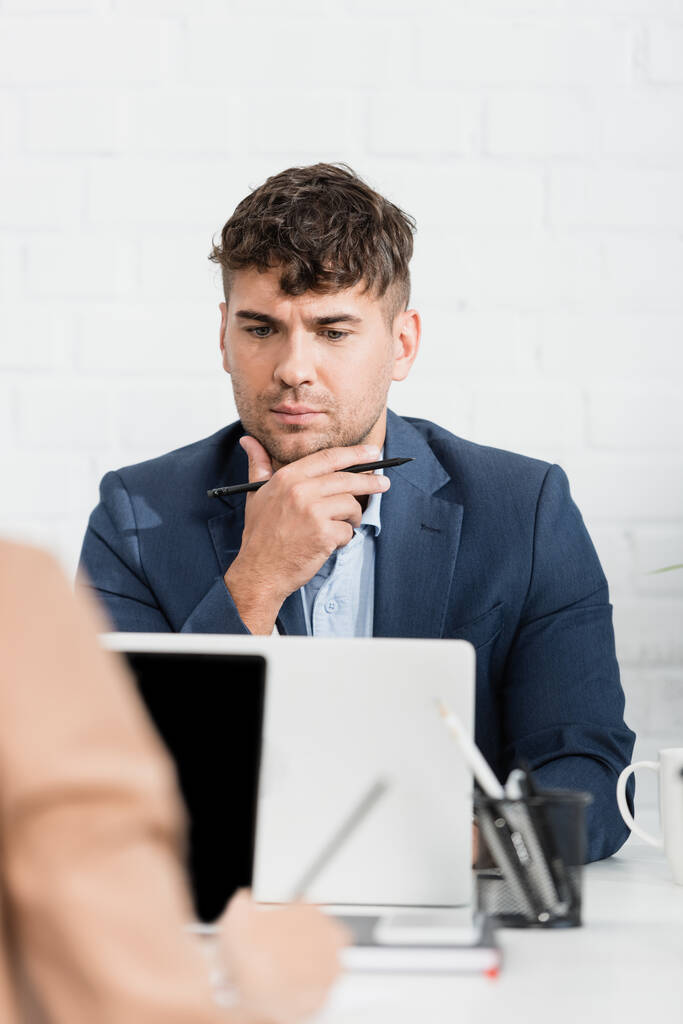 Empresário atencioso olhando para laptop, enquanto sentado à mesa no escritório em primeiro plano turvo - Foto, Imagem