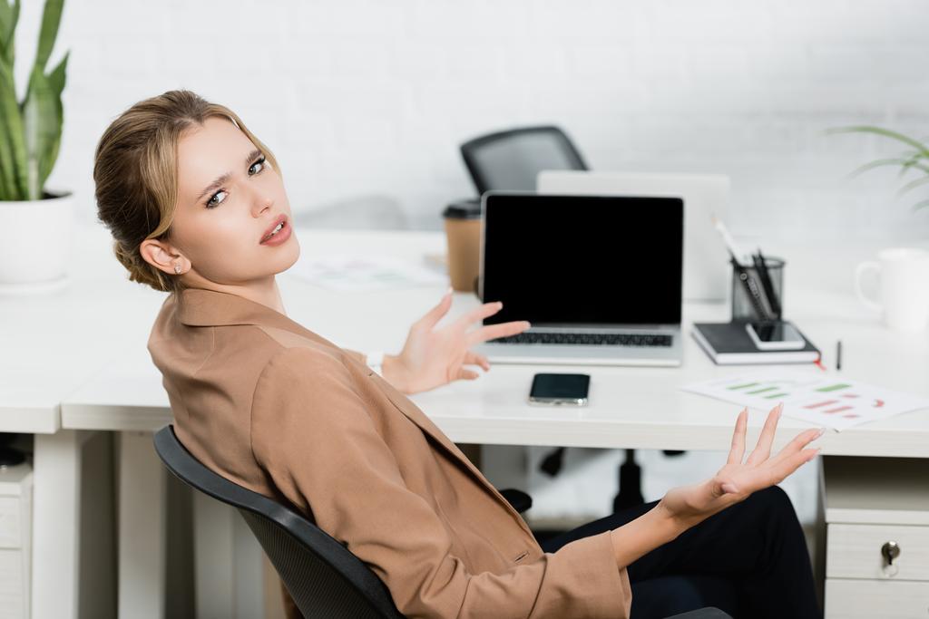 Irritierte weibliche Führungskraft blickt auf Kamera und gestikuliert, während sie am Arbeitsplatz mit digitalen Geräten auf verschwommenem Hintergrund sitzt - Foto, Bild