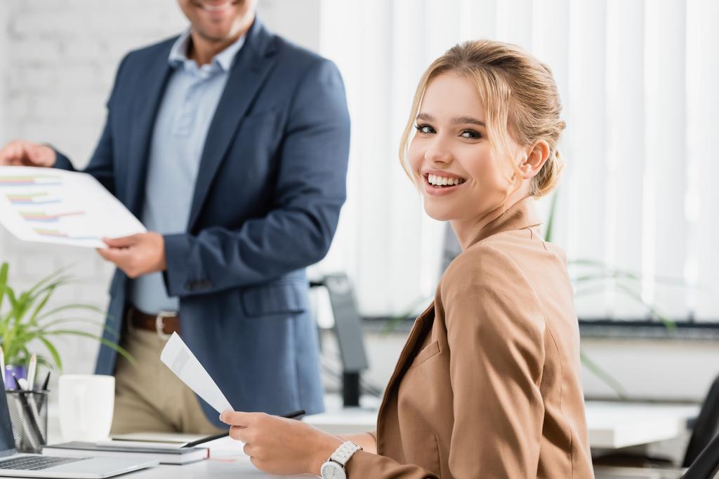 Glückliche Geschäftsfrau mit Dokument blickt in die Kamera, während sie am Arbeitsplatz mit verschwommenem Kollegen im Hintergrund sitzt - Foto, Bild