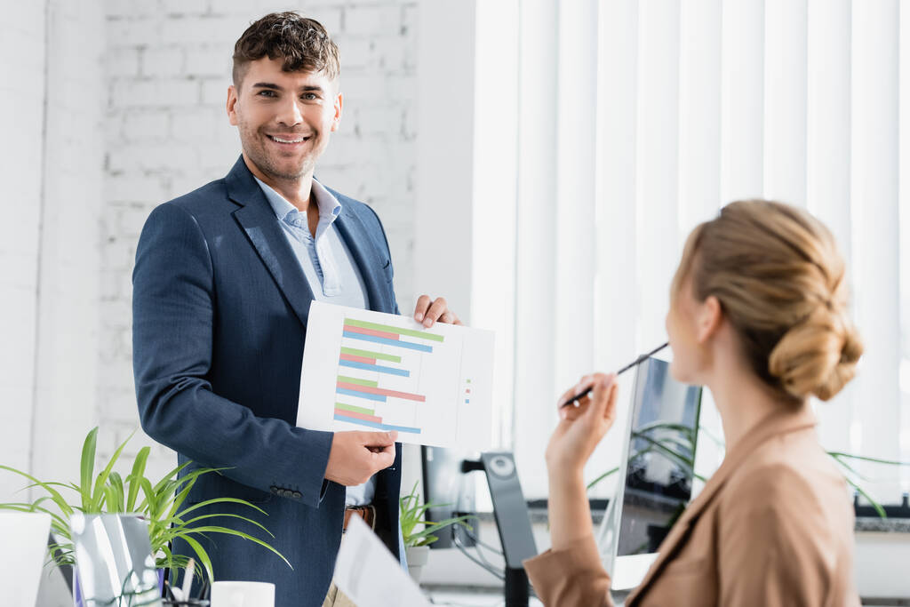 Hombre de negocios sonriente mirando a la cámara, mientras muestra una hoja de papel con gráficos cerca del lugar de trabajo en primer plano borroso - Foto, imagen