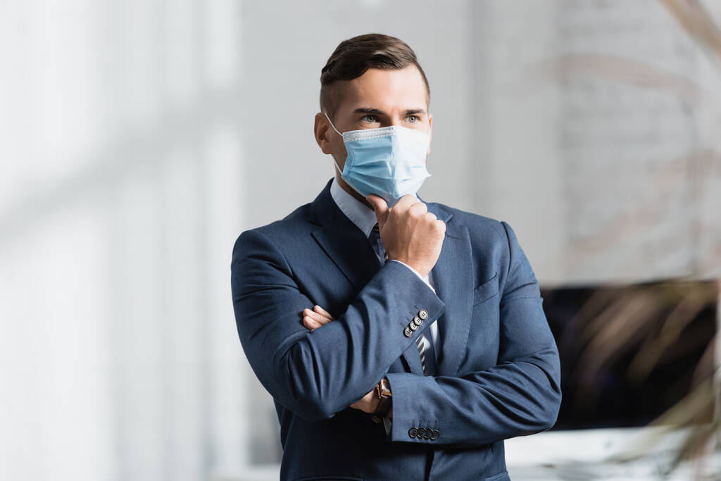 Продуманий бізнесмен в медичній масці, дивлячись вбік, стоячи в офісі на розмитому передньому плані
 - Фото, зображення