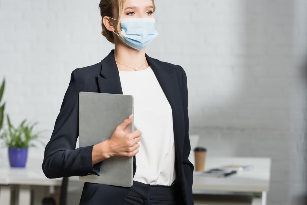 Жінка-виконавця в медичній масці з ноутбуком, дивлячись геть, стоячи в офісі на розмитому фоні
 - Фото, зображення