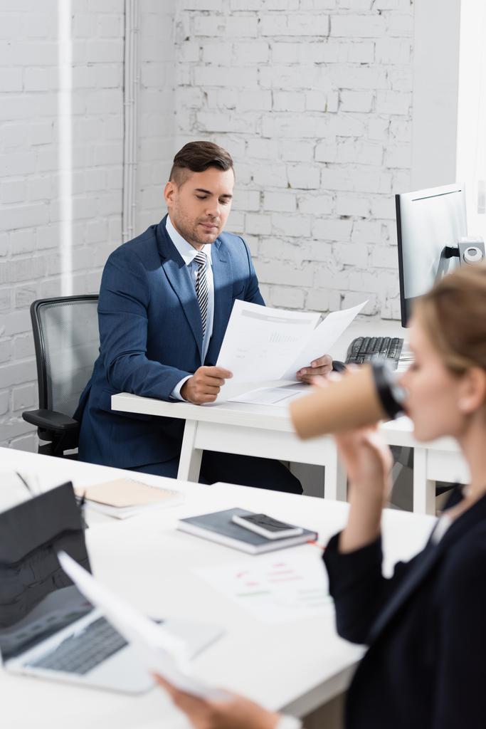 Geschäftsmann blickt auf Papierbögen, während er am Arbeitsplatz sitzt, mit verschwommener Kollegin im Vordergrund - Foto, Bild